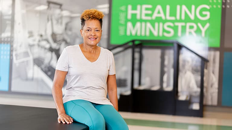 A Baptist Health Rehabilitation patient smiles at the camera while sitting in the therapy gym.