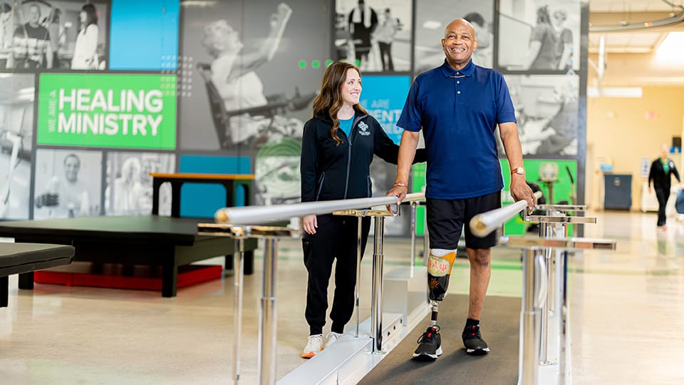 Man with amputated leg practices walking in between two bars alongside therapist.