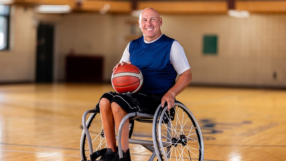 A man in a wheelchair on a basketball court holding a basketball smiles at the camera.