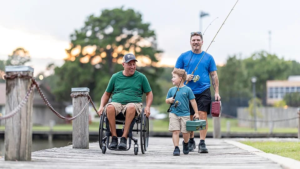 Man in wheelchair with son and grandson carry fishing equipment
