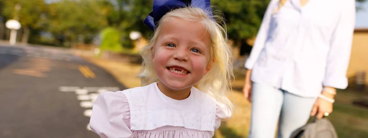 Smiling young girl with blonde hair and a large blue bow, wearing a light pink dress, standing outdoors with an adult nearby.