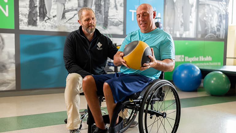 Man in wheelchair works out with exercise ball alongside therapist.
