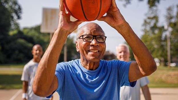 Smiling older man in glasses wearing a blue shirt holds a basketball overhead while playing with friends on an outdoor court.