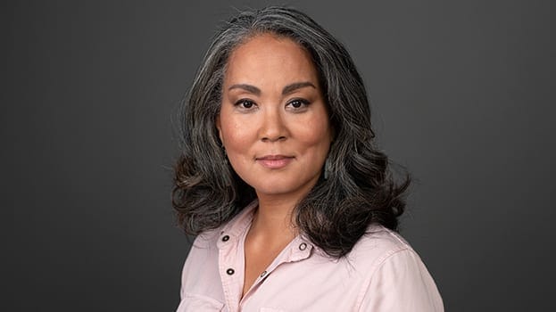 A woman with long, wavy salt-and-pepper hair wearing a light pink button-up shirt, looking confidently at the camera against a plain dark background.